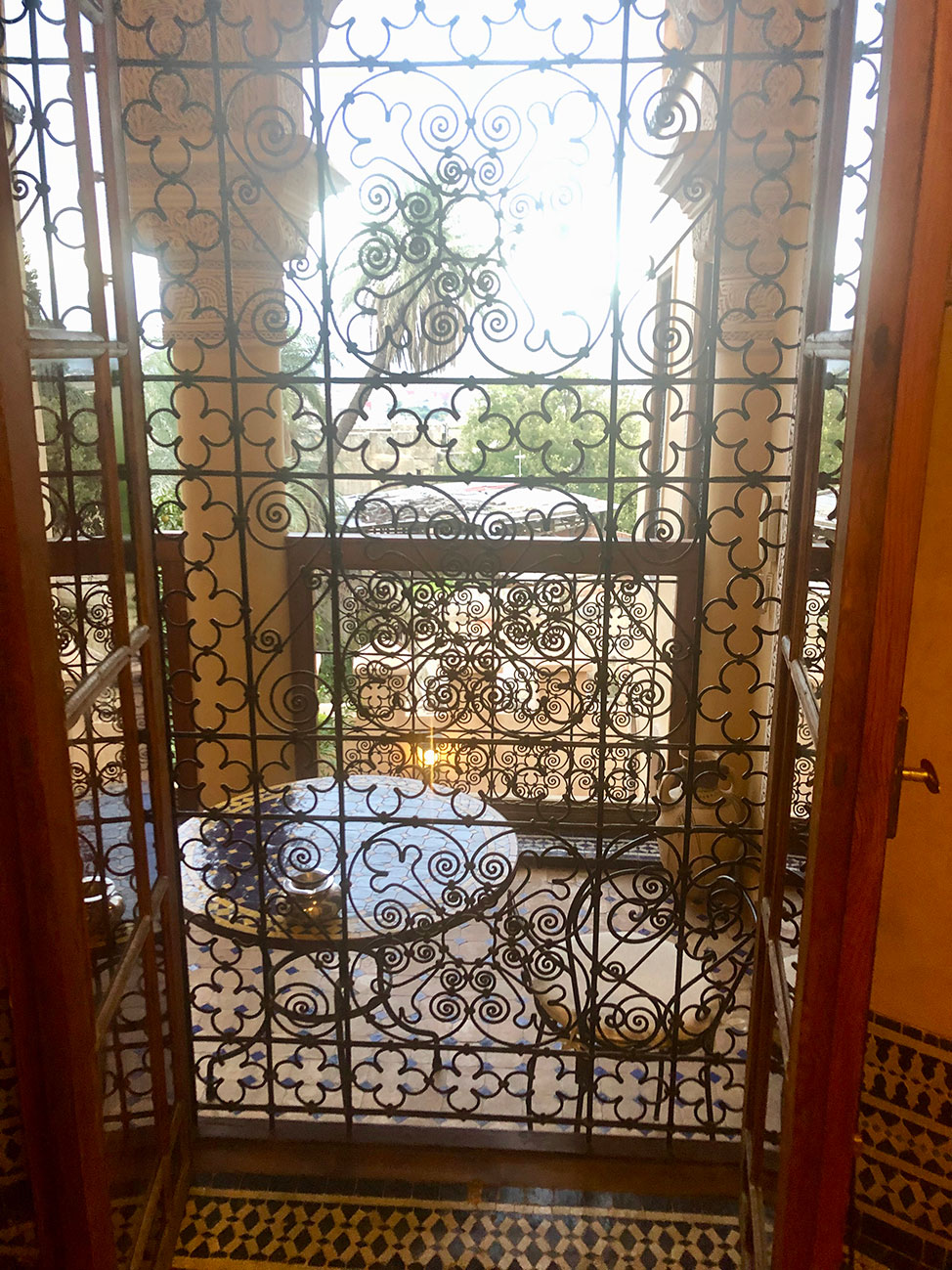 Looking onto a sunny terrace from behind a window covered by decorative wrought iron.