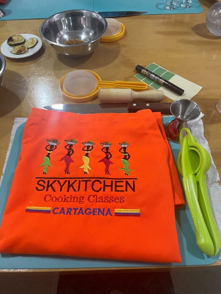 Bright orange apron with the words Sky Kitchen Cooking Classes - Cartagena surrounded by cooking tools.