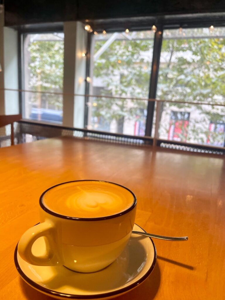 A cappuccino sits on a table in a cafe with a window view of the street.